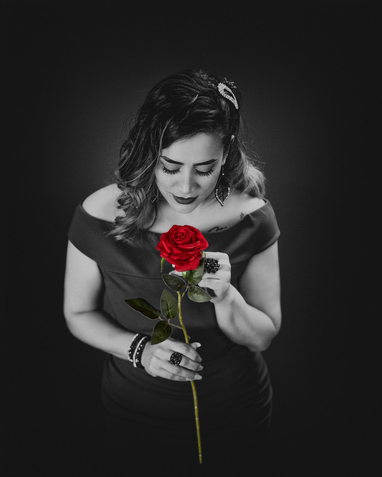 Monochromatic portrait highlighting red rose against black background with off-shoulder dress.