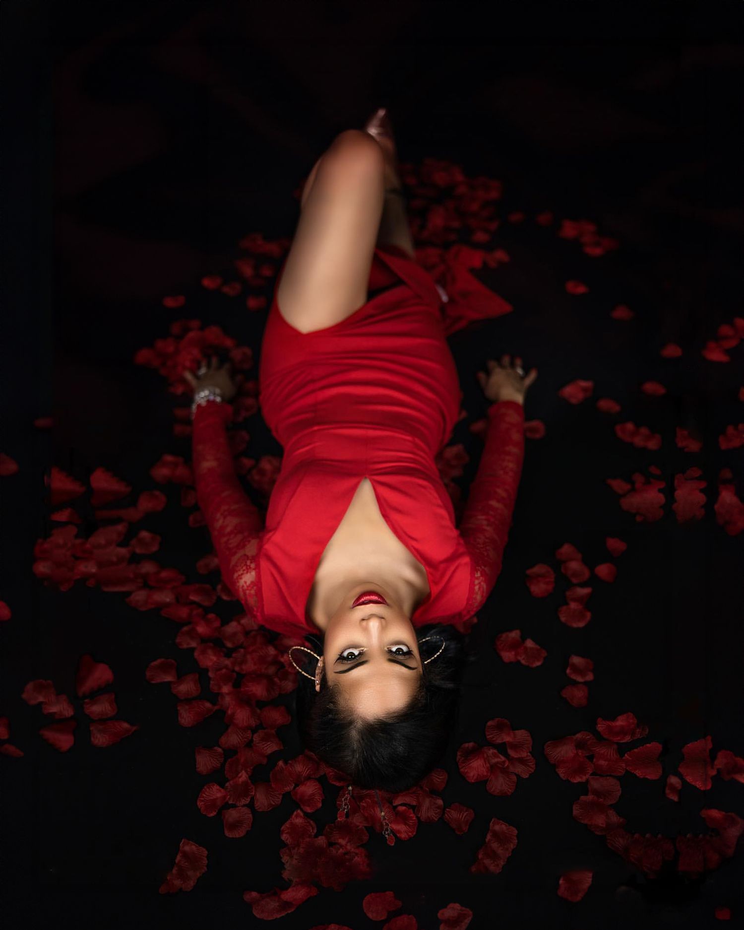Dramatic overhead portrait in red dress surrounded by rose petals on dark background.