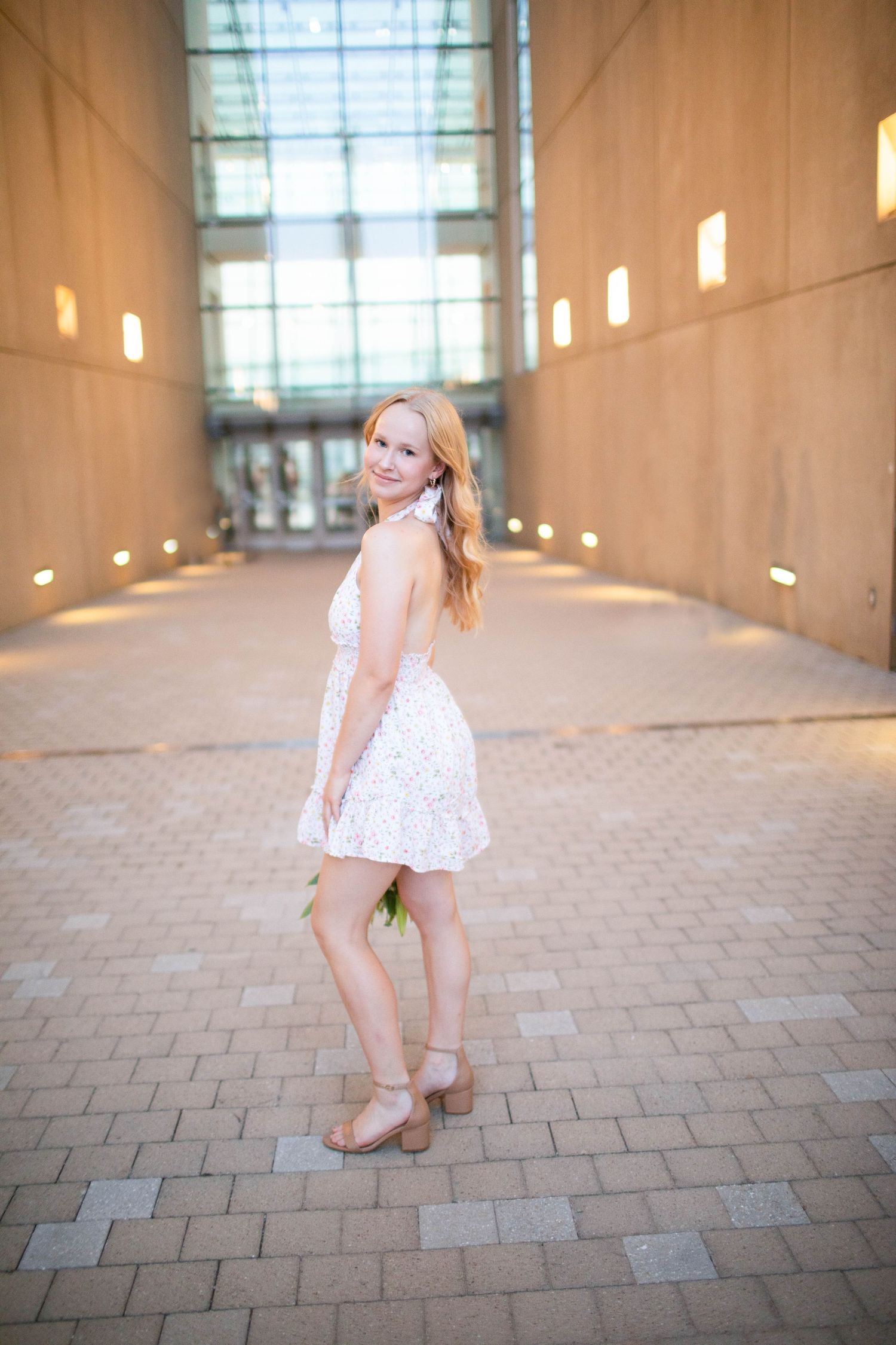 Young woman in white dress poses in sunlit hallway with brick floor.