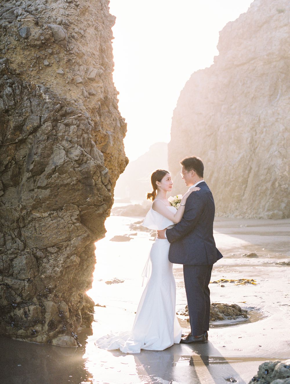 Bride and groom embracing between rocky cliffs on beach at sunset.