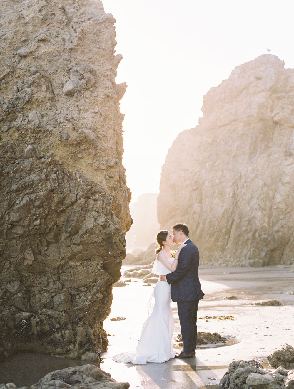 Newlyweds share intimate moment between coastal rock formations.