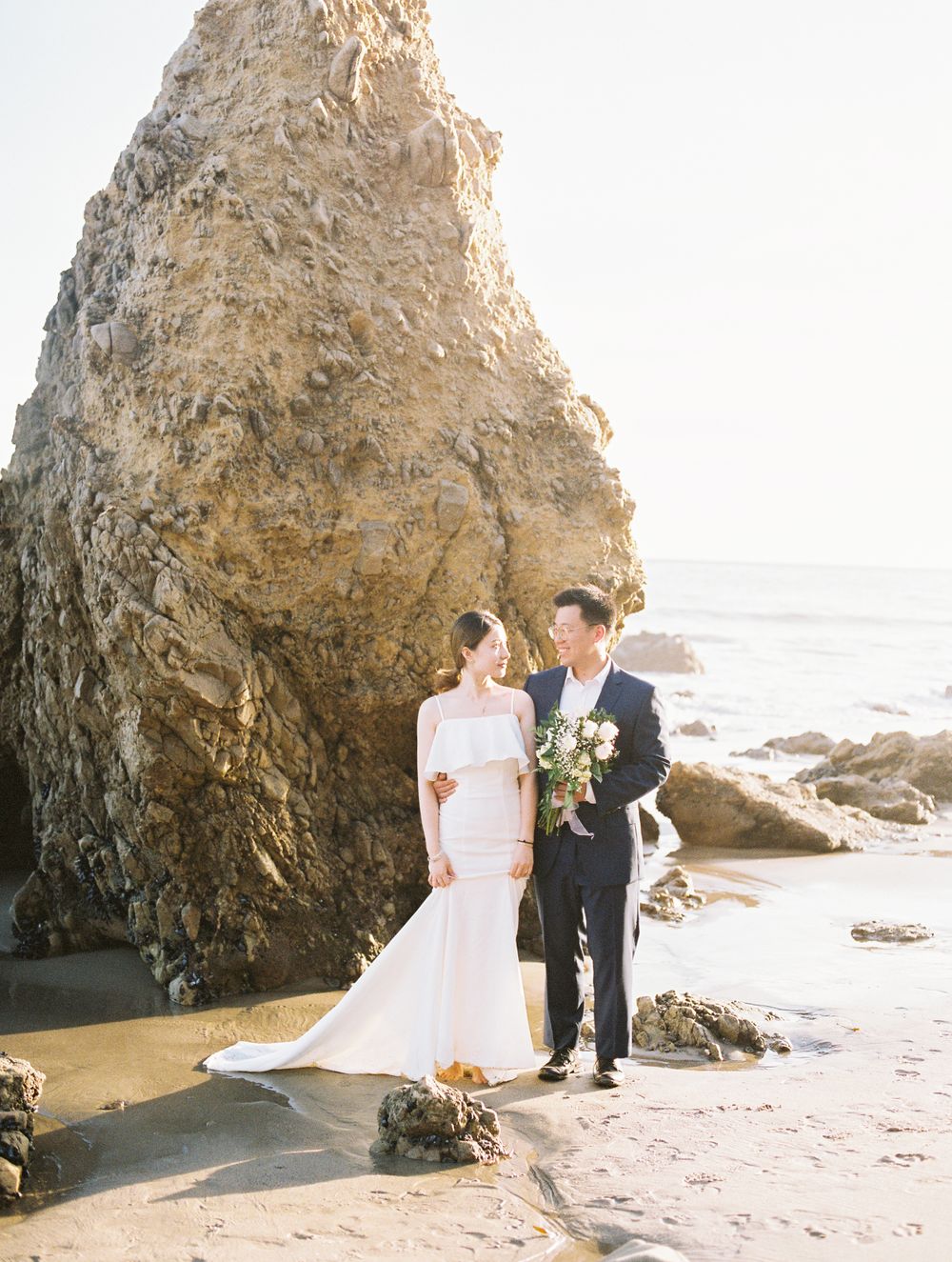 Wedding couple poses by large coastal rock formation on beach.