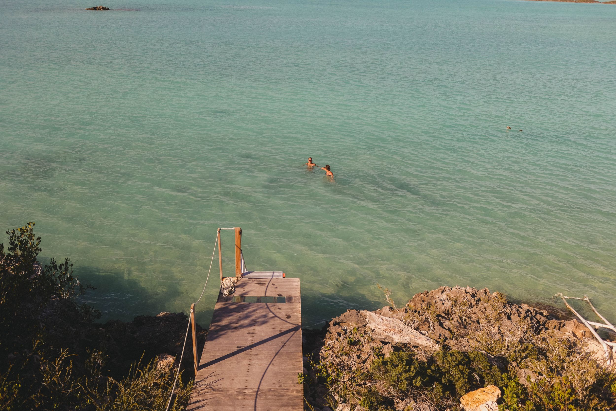 Turquoise ocean waters meet a wooden boardwalk along a rocky coastline on a sunny day.