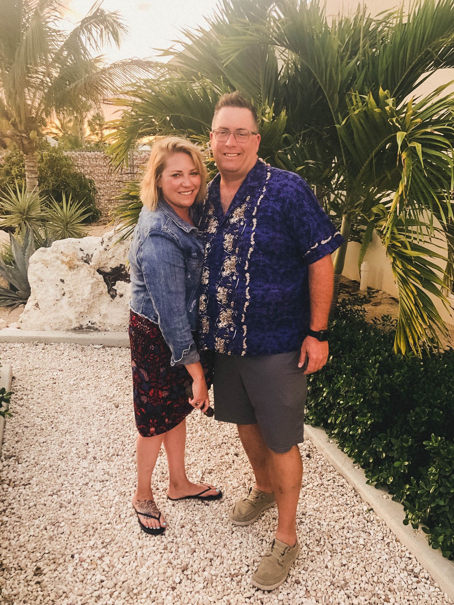 Couple poses together on a tropical pathway surrounded by palm trees and white stones.