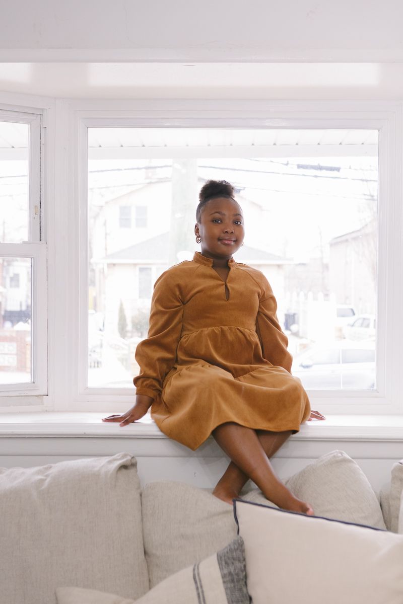 a full body view of a little girl sitting by the window at home in Queens during a family session by NYC family photos