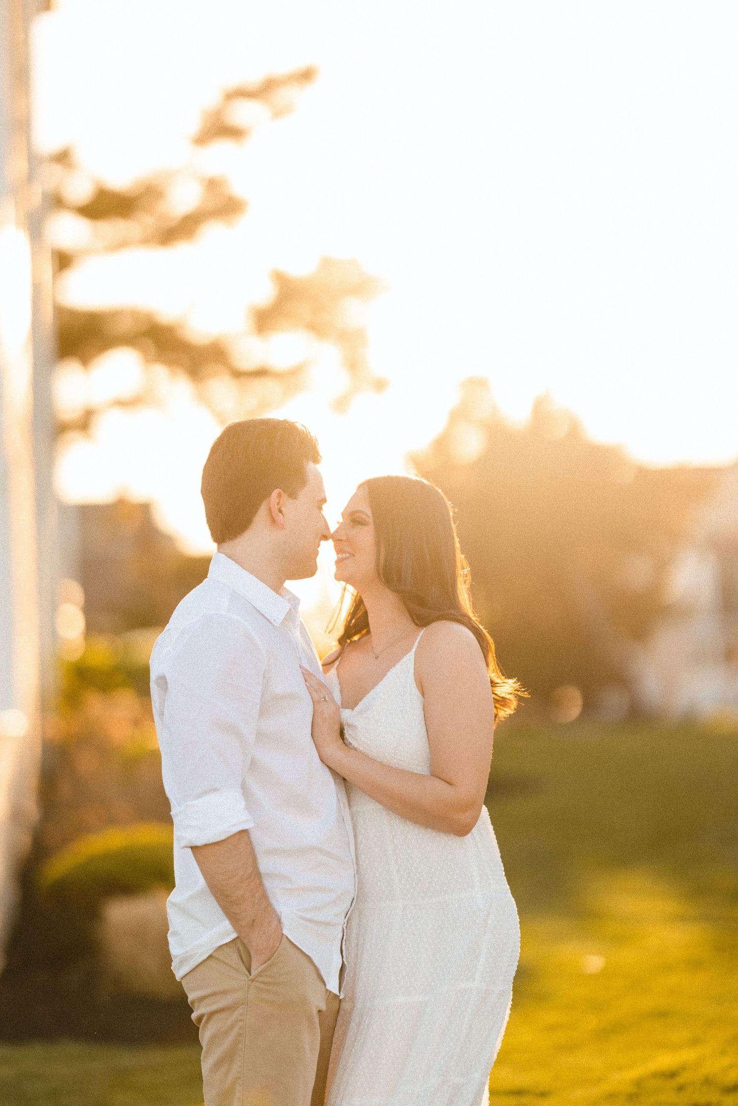 Couple almost kissing at golden hour sunset, woman in white sundress and man in white shirt embracing outdoors.