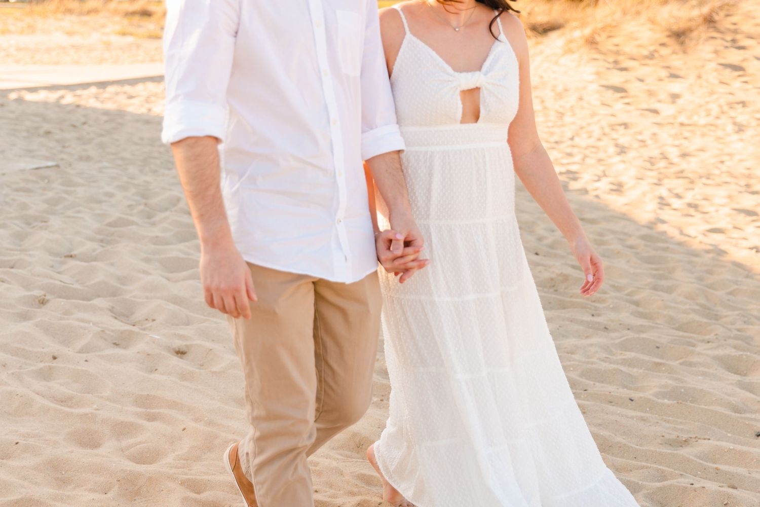 Couple walking hand in hand on a sandy beach, woman in white sundress and man in khaki pants.