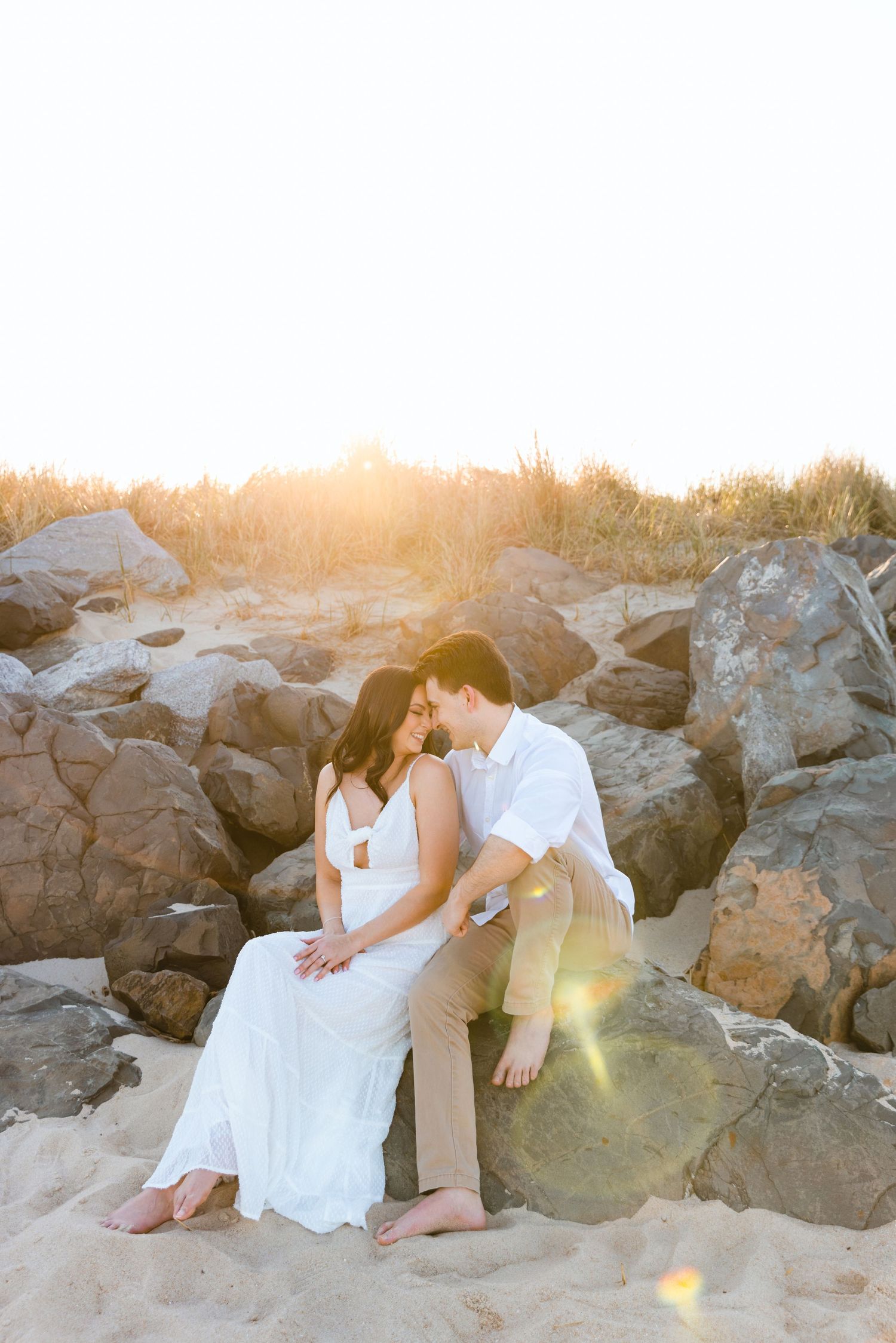 Couple sitting on beach rocks at golden sunset, woman in white dress and man in white shirt sharing a romantic moment.