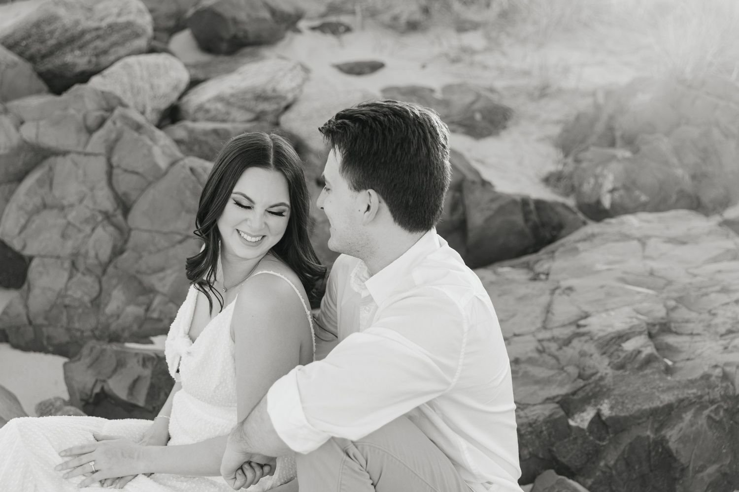 Black and white photo of a smiling couple sitting together in front of large rocks on the beach.