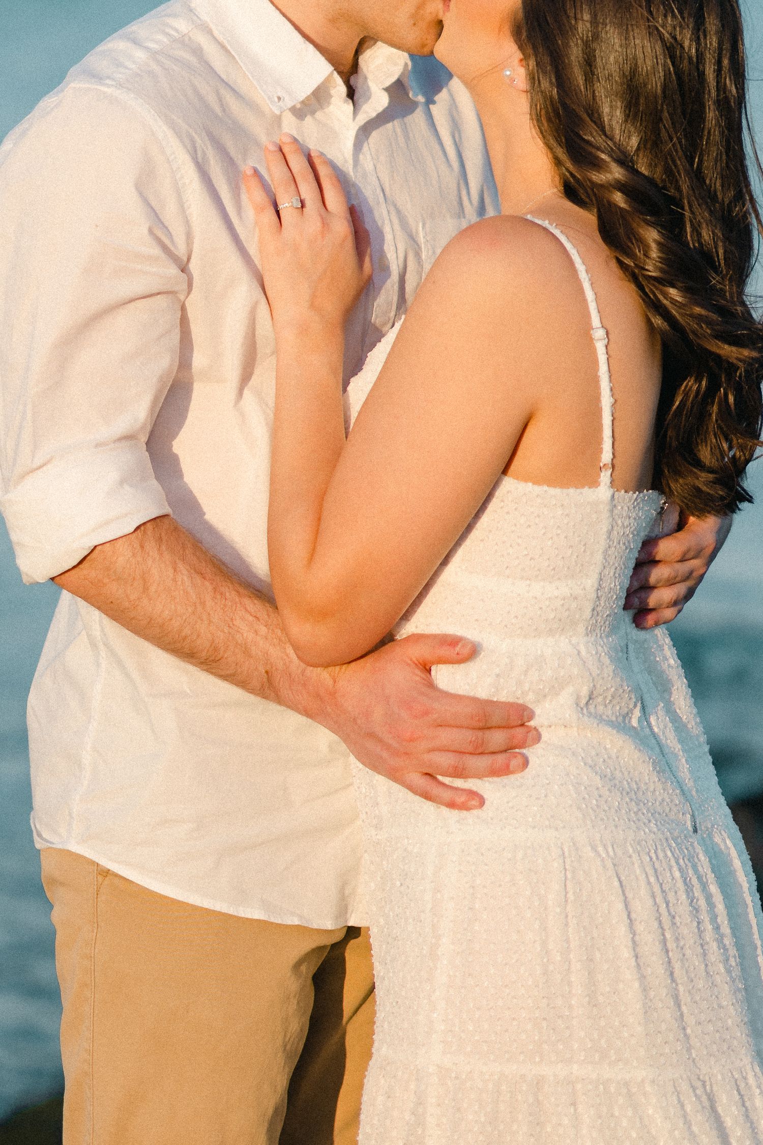 Close-up of couple kissing and embracing, woman in white lace dress with man's hand on her waist by the sea.