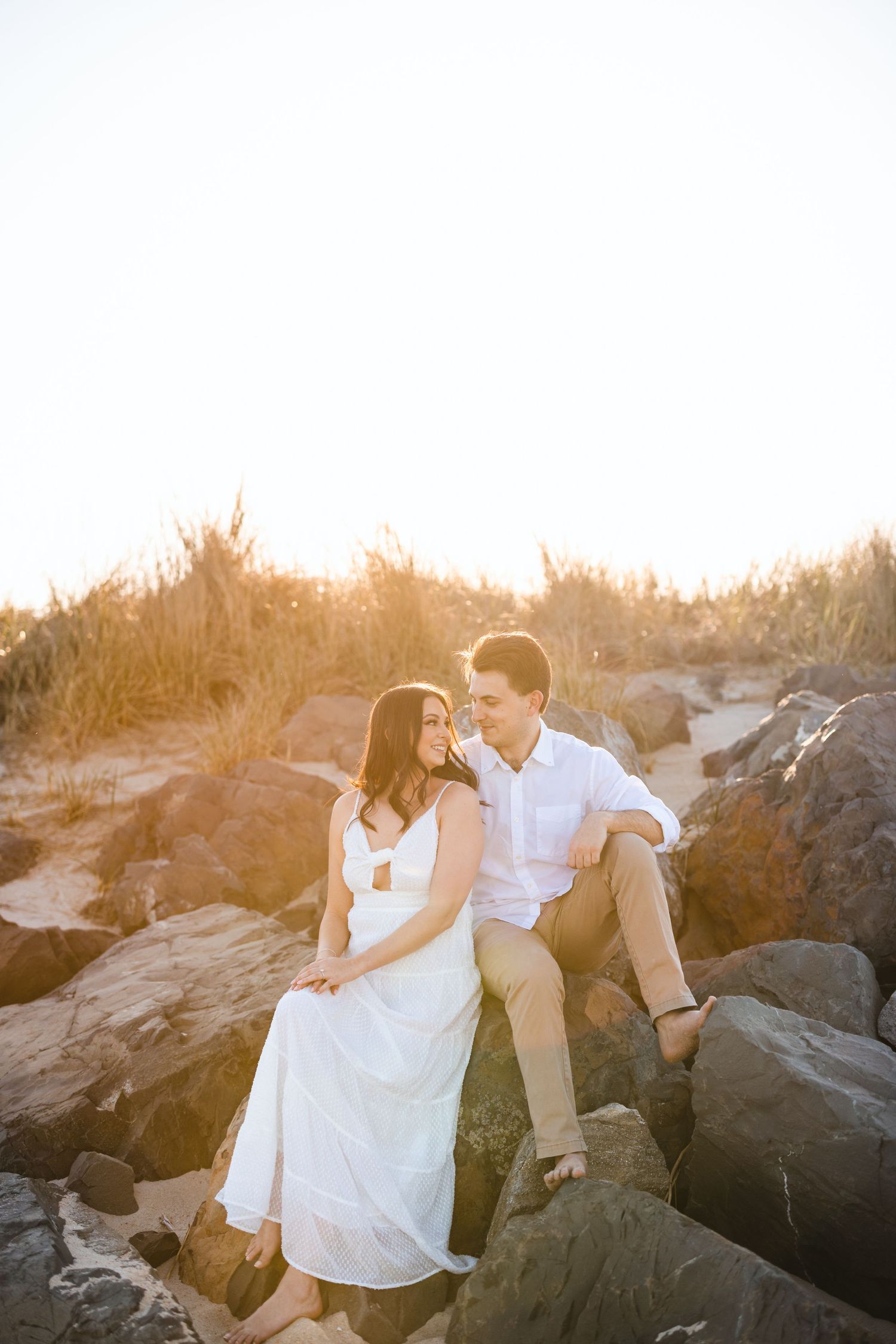 Couple sitting together on large rocks at golden hour, woman in white dress smiling at man in white shirt.