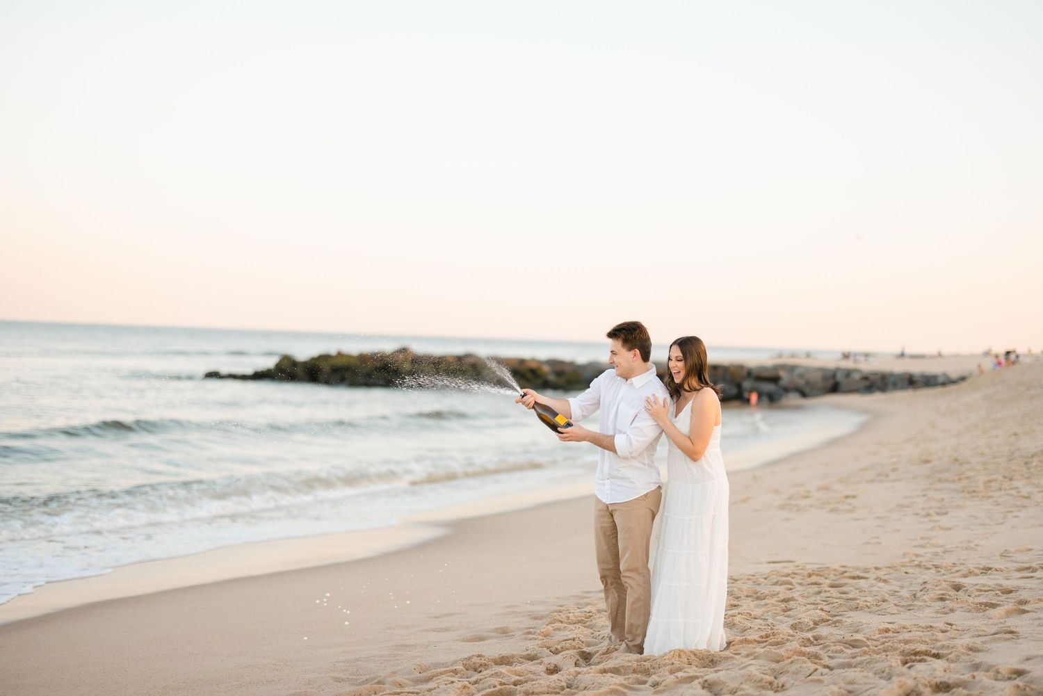 Couple standing on sandy beach at sunset, man holding champagne bottle with ocean waves in background.