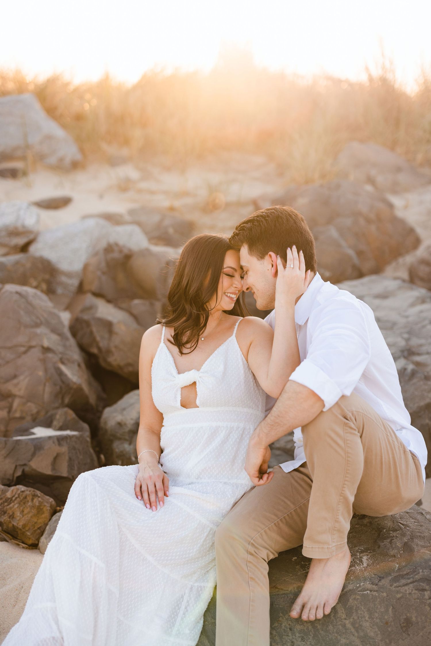 Couple sharing a tender forehead-to-forehead moment on beach rocks at warm golden sunset, woman in white dress.