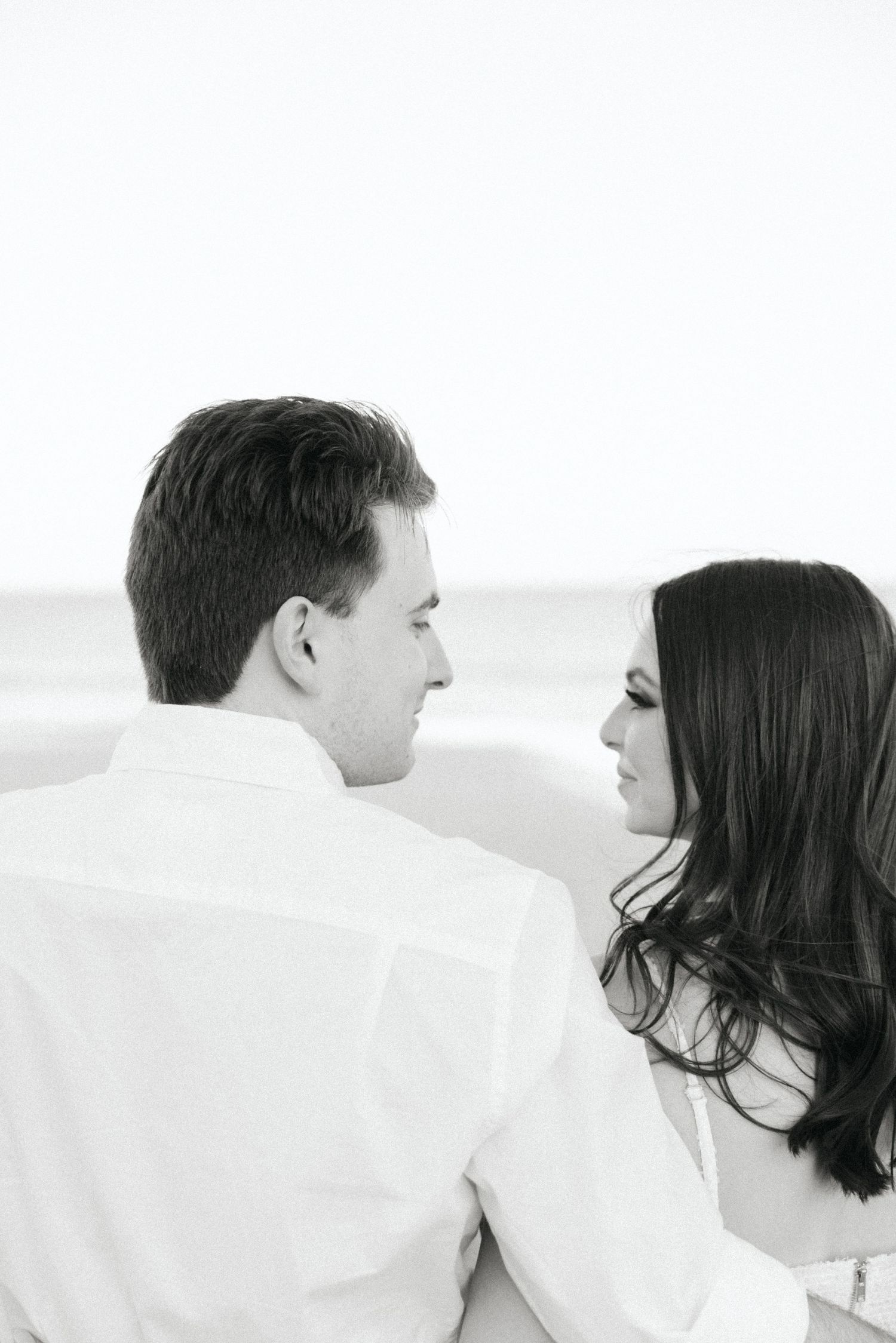 Black and white photo of an engaged couple gazing at each other near the ocean beach shoreline.