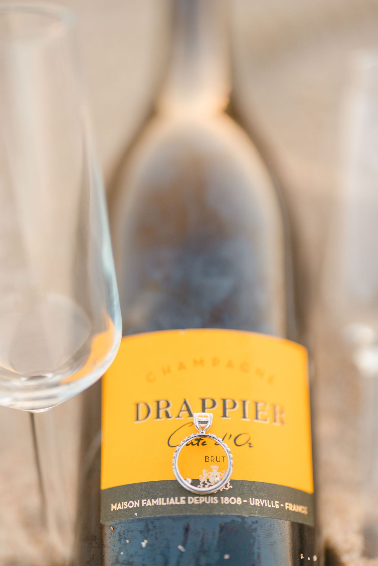 Drappier Carte d'Or Brut Champagne bottle with engagement ring and flute glasses on sandy beach surface.