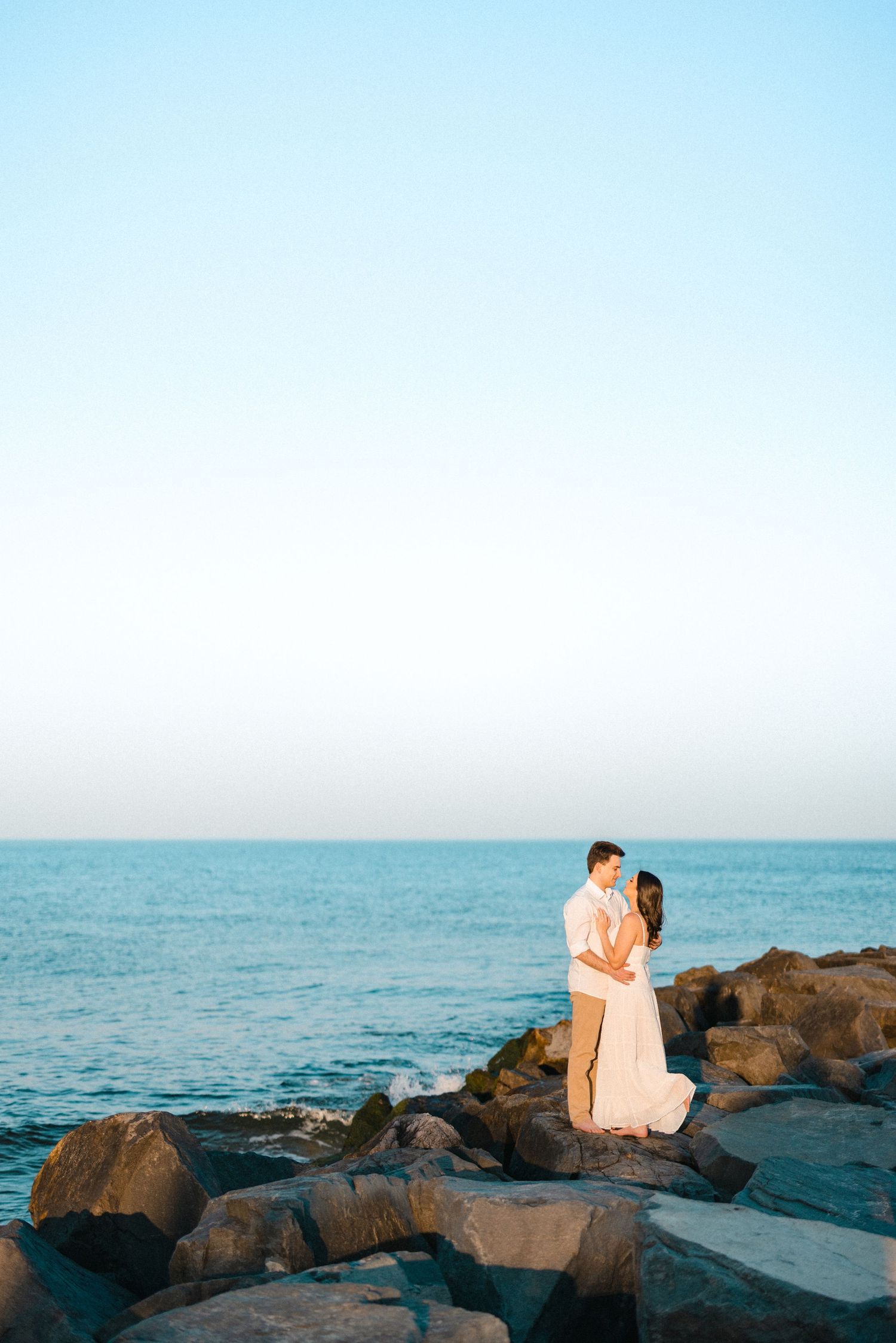 Romantic couple embracing on rocky jetty with calm blue ocean and clear sky in the background.