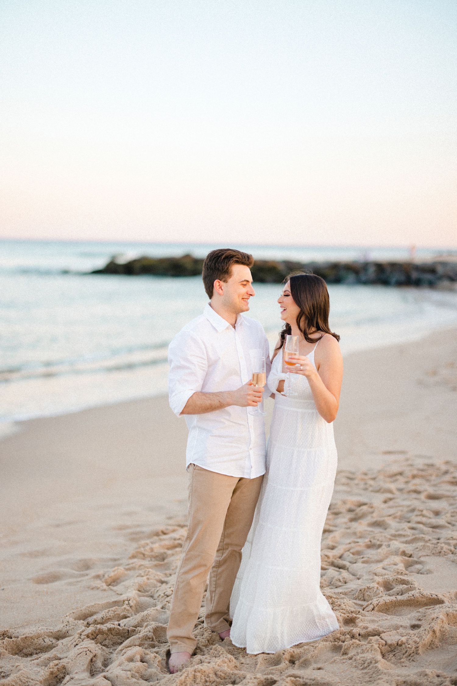 Happy couple laughing and holding champagne glasses on a sandy beach at pastel sunset near a rocky jetty.