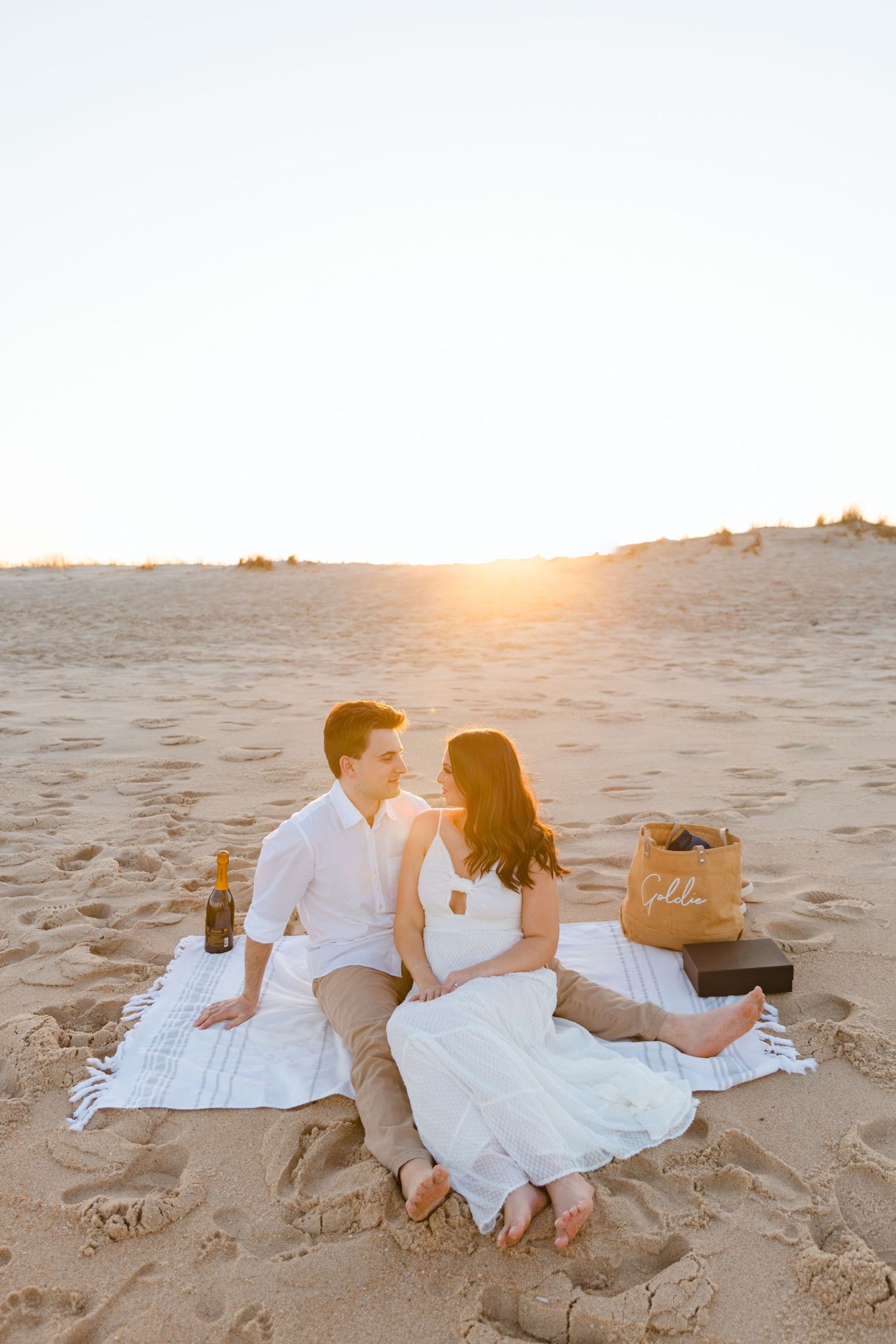 Engaged couple sitting on a blanket during a golden sunset beach picnic with champagne and a wicker bag.