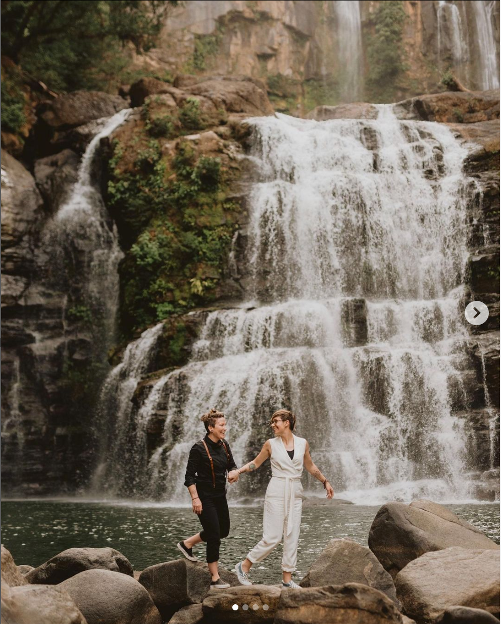A couple standing on rocks near a majestic cascading waterfall surrounded by lush forest.