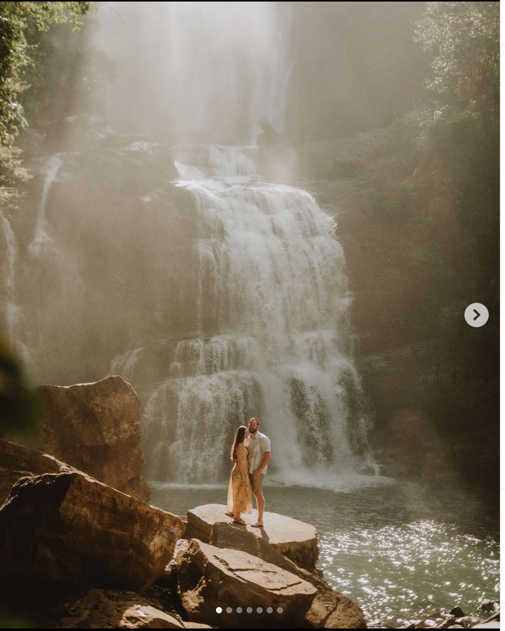 A couple embraces on a rocky outcrop with a misty waterfall cascading behind them.