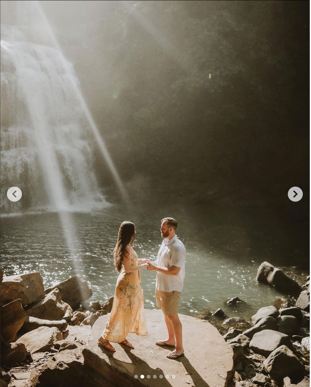 Two people share an intimate moment in front of a sunlit waterfall with rays streaming through the mist.