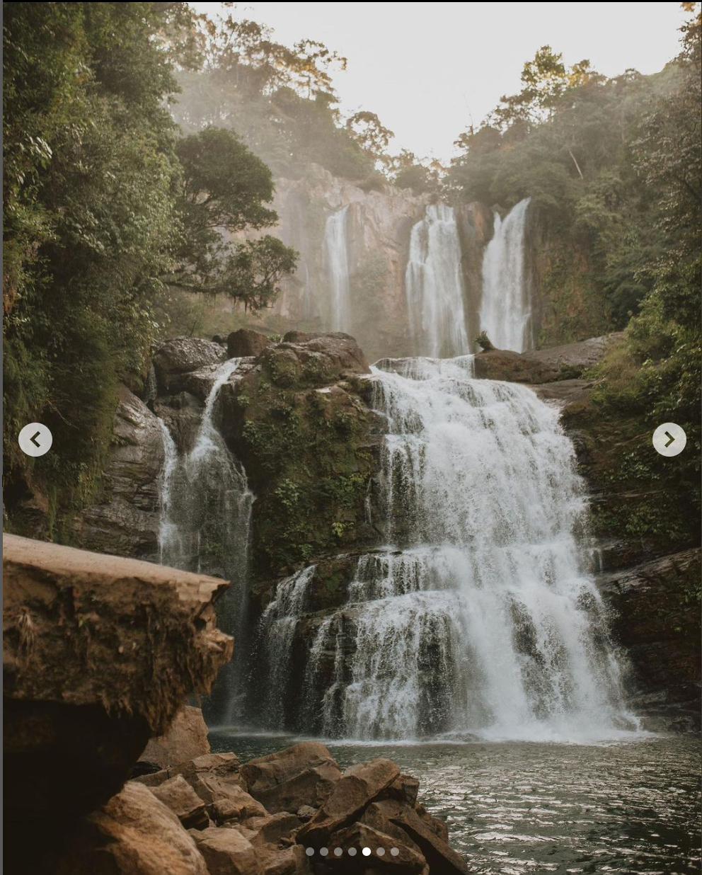 A multi-tiered waterfall flows dramatically over rocky cliffs surrounded by green forest.