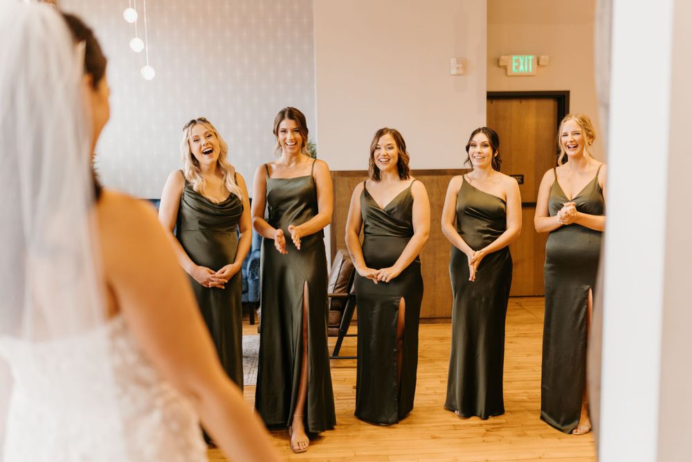 Bridesmaids in dark olive satin gowns reacting with joy during first look reveal of bride.