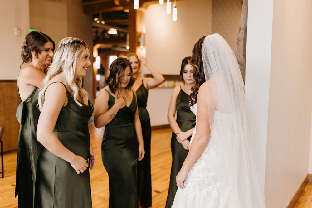 Bridesmaids in dark olive gowns gathering around bride in lace wedding dress during first look.