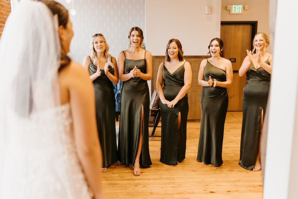 Excited bridesmaids in dark green satin dresses clapping during emotional first look with bride.
