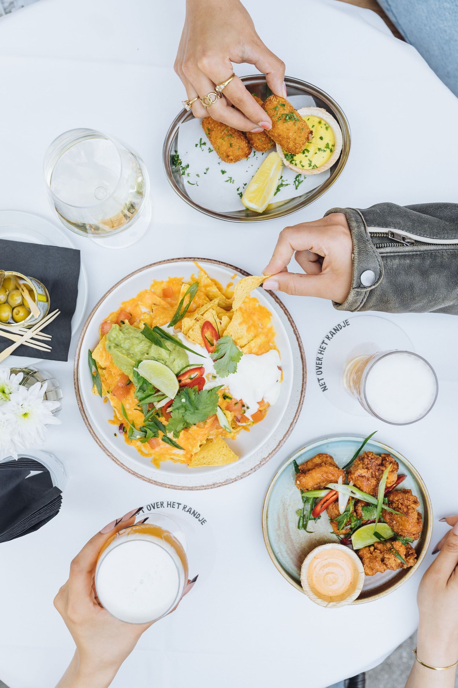Overhead view of colorful shared plates with drinks on white tablecloth at restaurant.