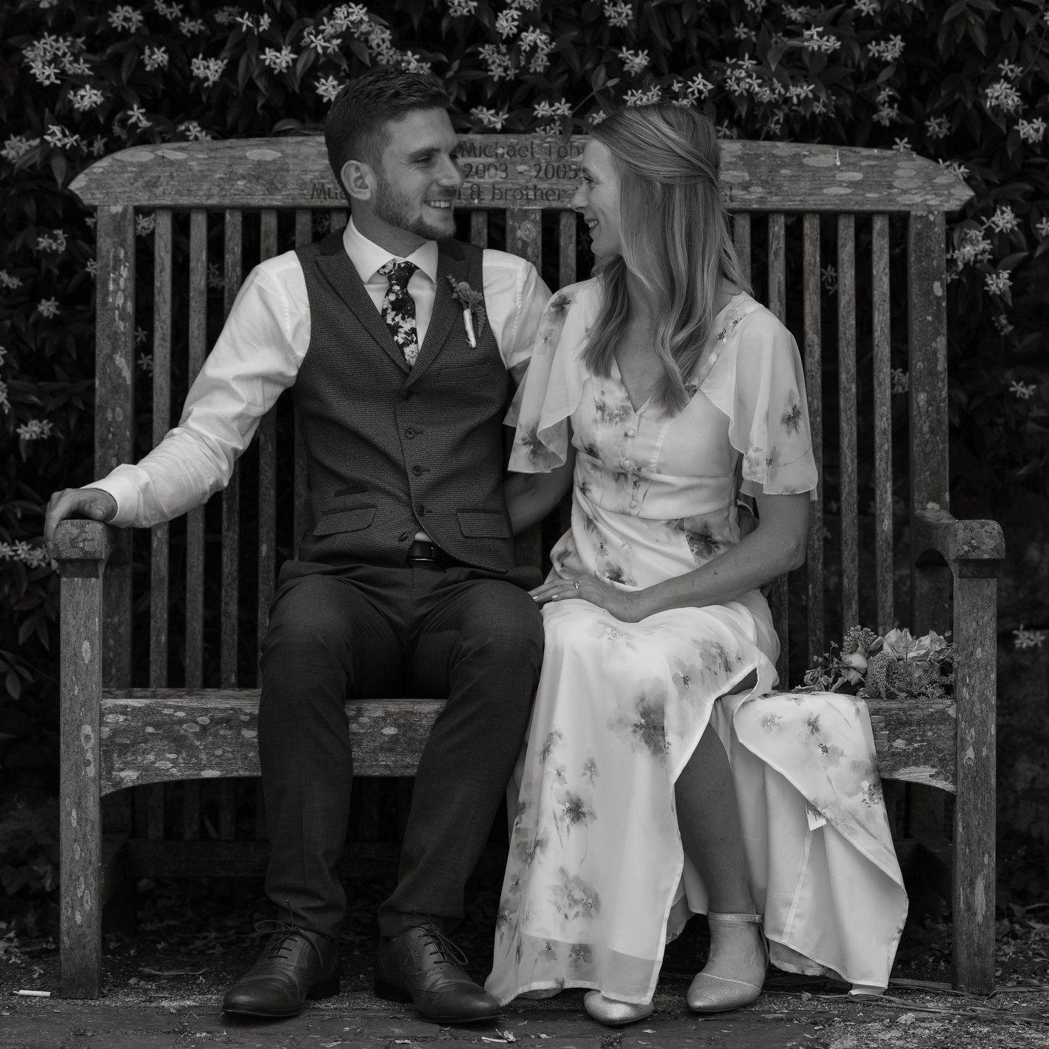 A couple sitting together on a wooden garden bench in formal attire.