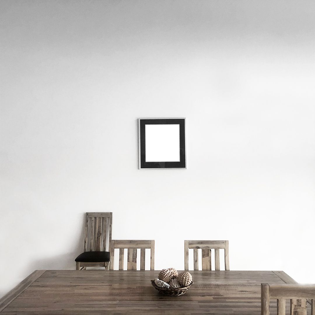 Minimalist dining room with empty black frame on white wall above wooden table and chairs.