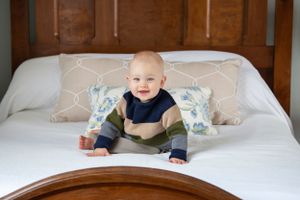 Happy baby wearing navy shirt sits on white bed with floral pillows.