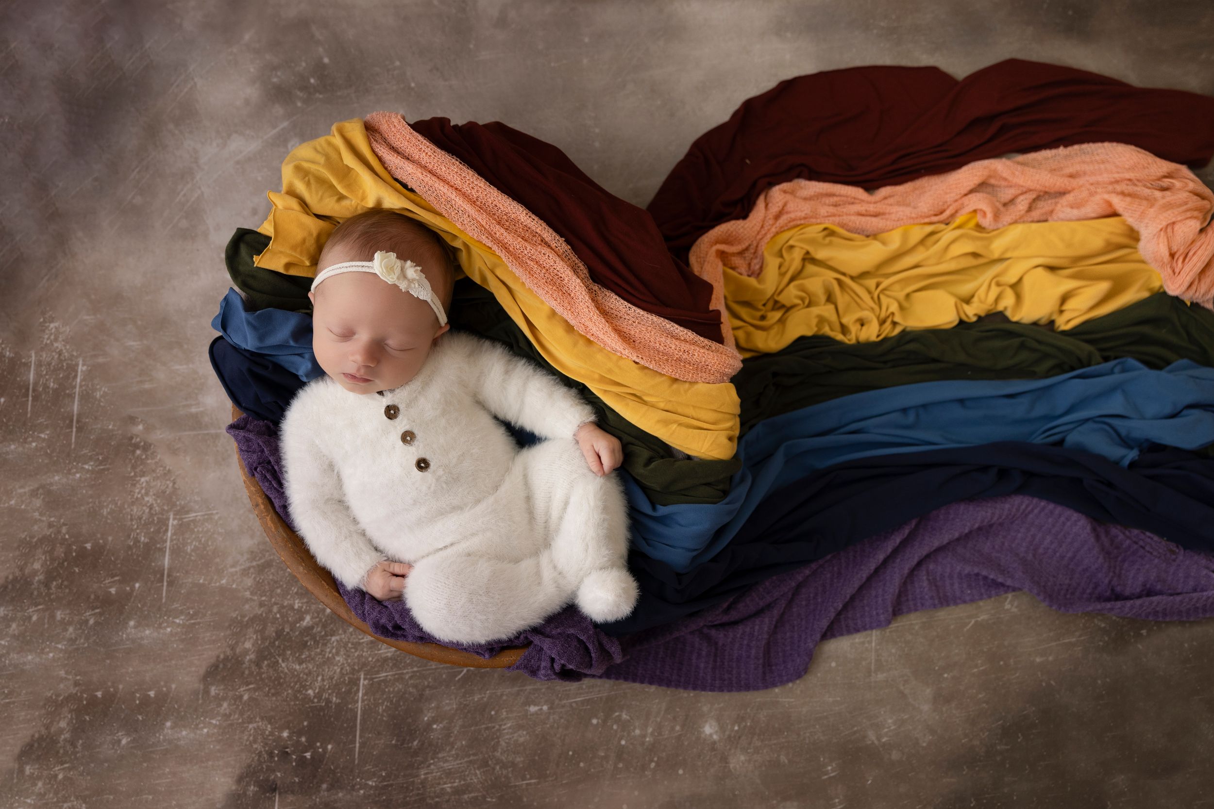 Newborn wrapped in white sleeps on rainbow colored fabric layers.