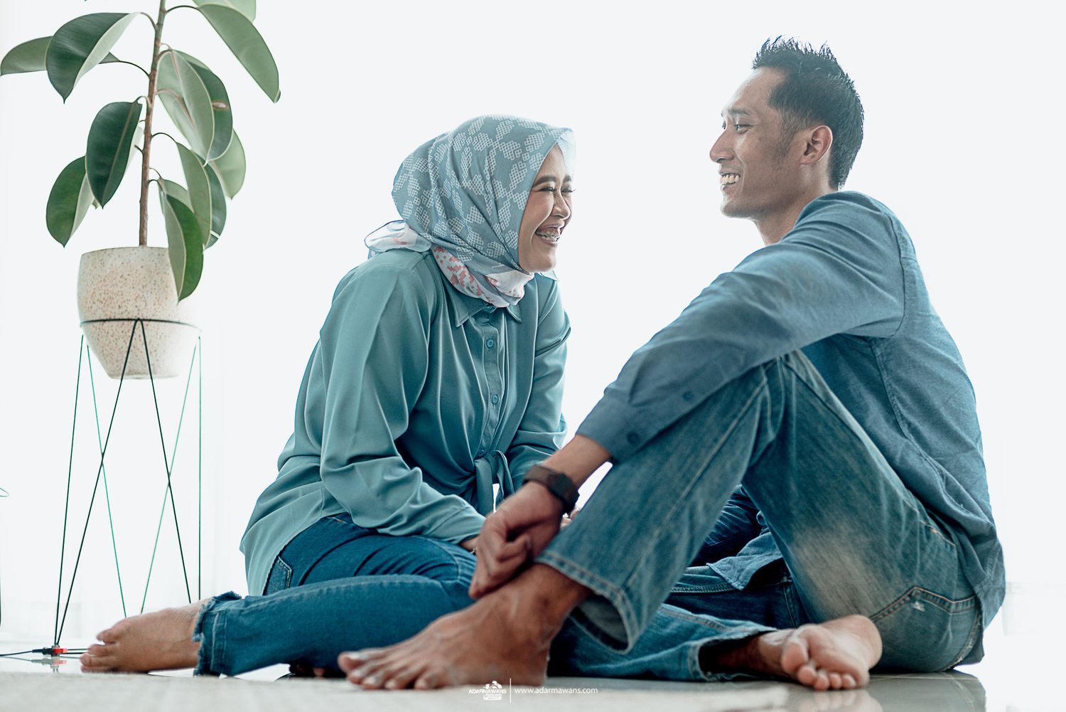 A couple in matching outfits sits cross-legged on the floor near a potted plant.