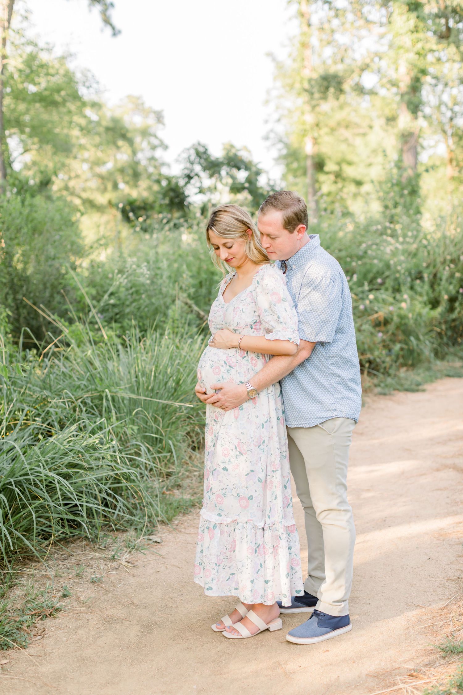 A sweet embrace between expecting parents on a dirt trail surrounded by tall grass.