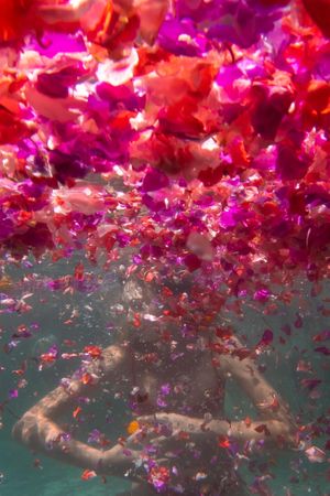 A woman embraces herself underwater surrounded by flowers