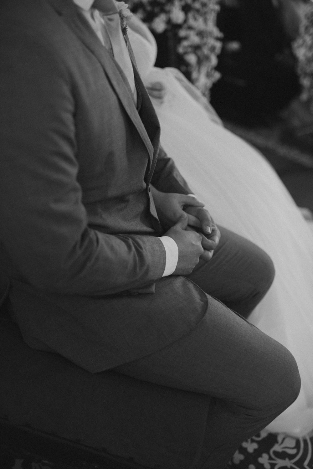 A person in a formal grey suit sits with clasped hands during a wedding ceremony.