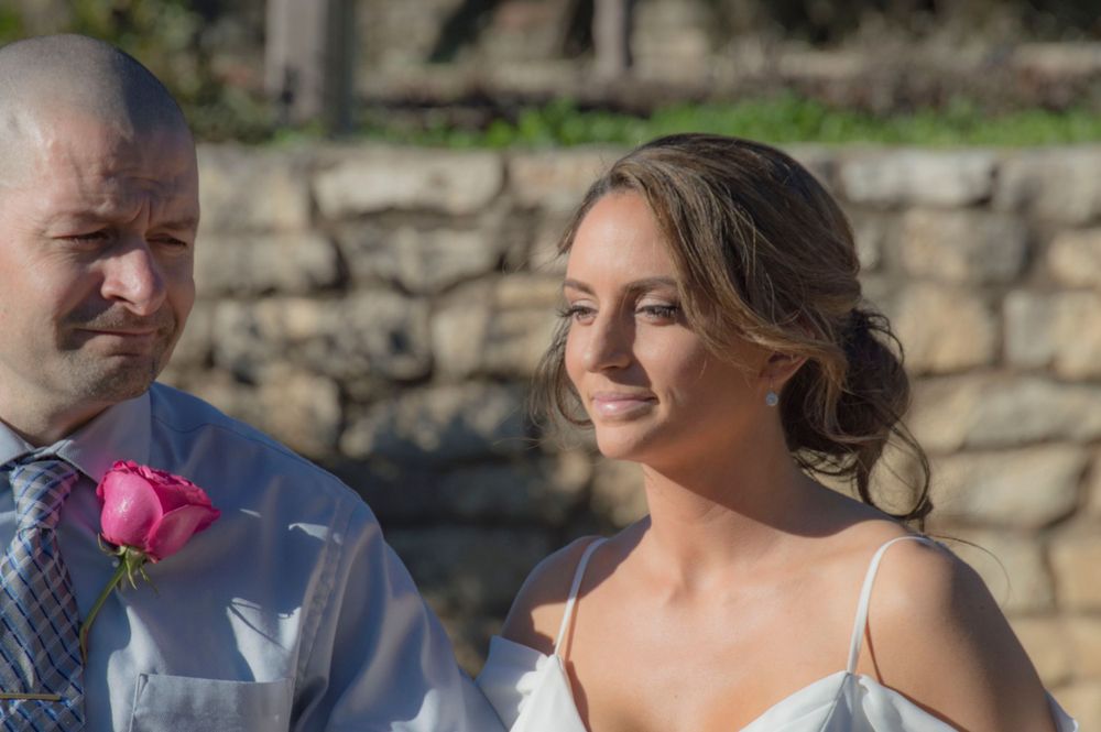 Bride smiling warmly during outdoor wedding ceremony with stone wall background and pink boutonniere visible.