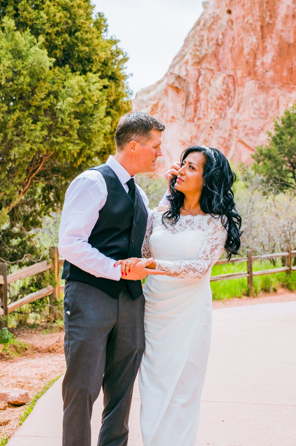 Bride in lace gown and groom in vest holding hands on path with red rock formations behind.