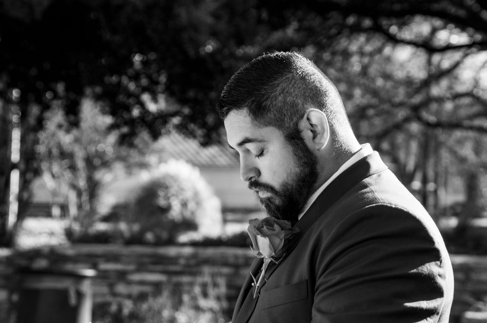 Black and white portrait of bearded groom in suit looking down thoughtfully in outdoor garden setting.