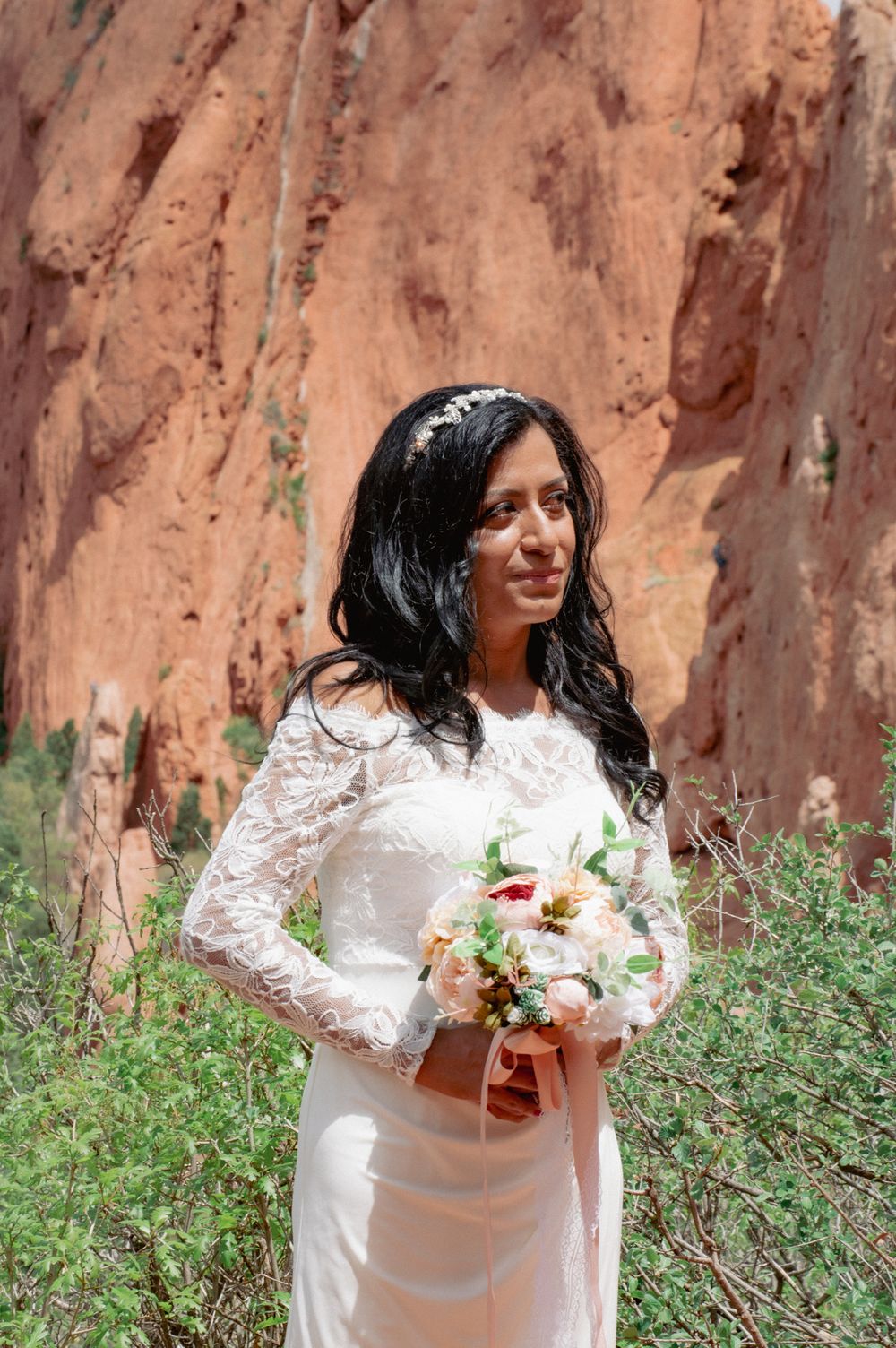 A bride in a white lace long-sleeve gown holds a bouquet in front of red rock formations.