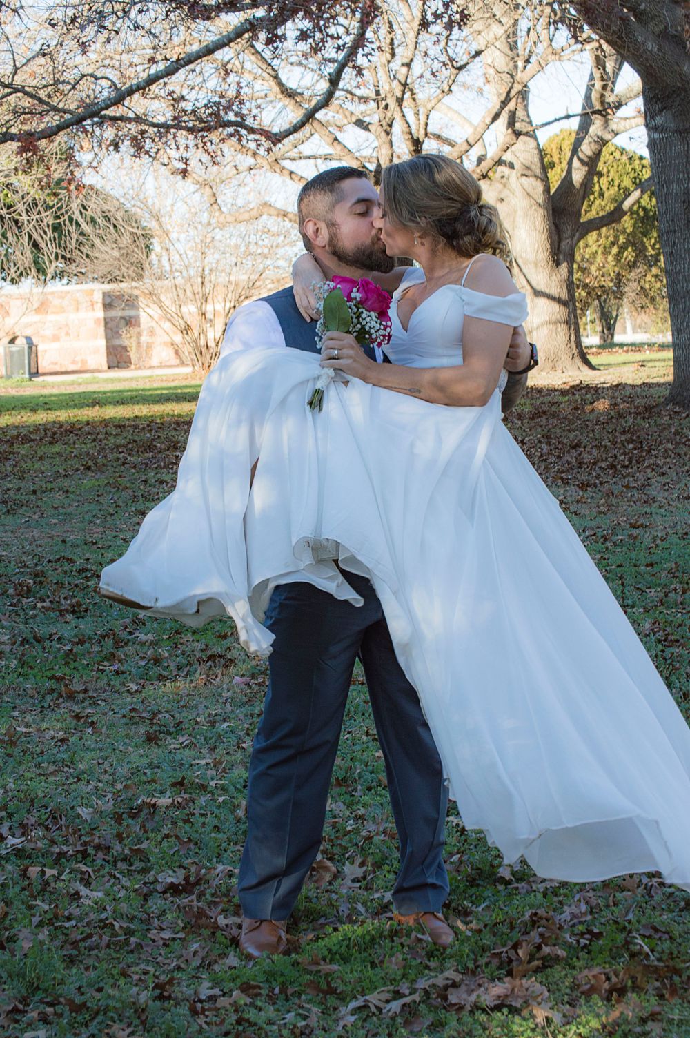 Bearded groom lifting bride in white gown for a kiss outdoors among autumn trees in park.