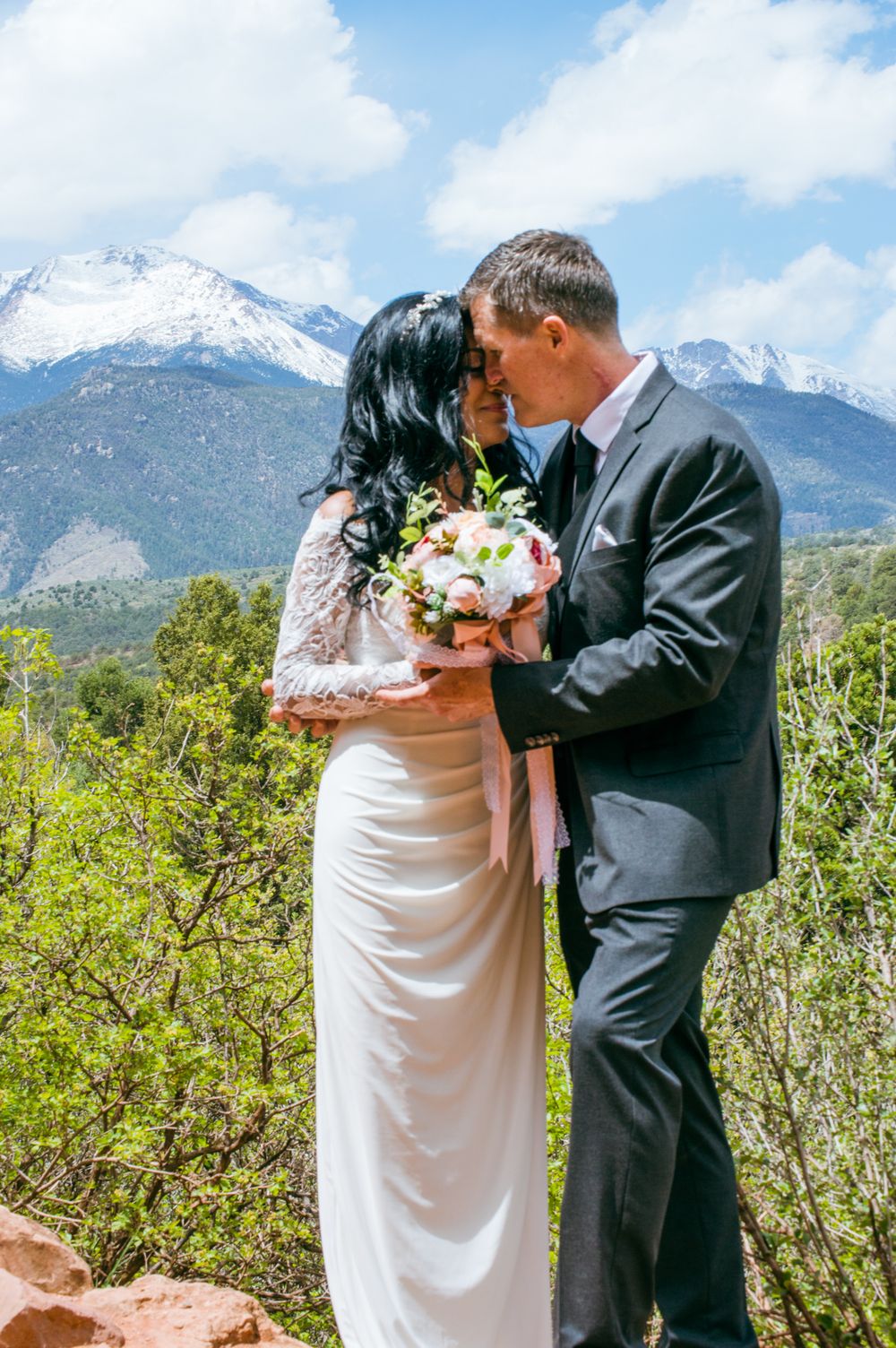 Couple kissing at outdoor wedding with snow-capped Pikes Peak mountains and lush green trees in background.