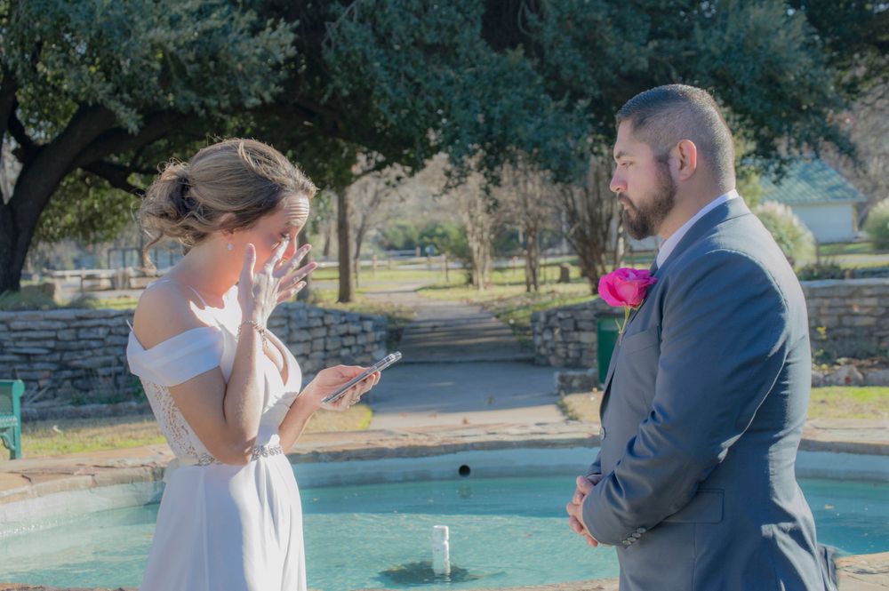 Bride reading wedding vows from card while emotional, facing bearded groom near fountain in sunny park.