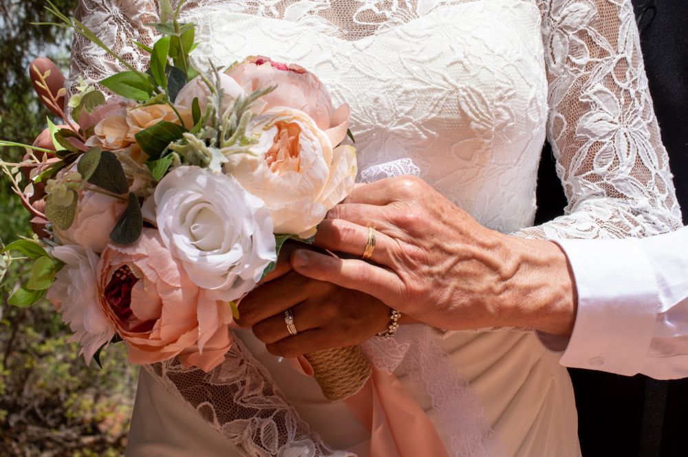 Close-up of bride and groom hands holding a pastel floral bouquet with wedding rings visible.