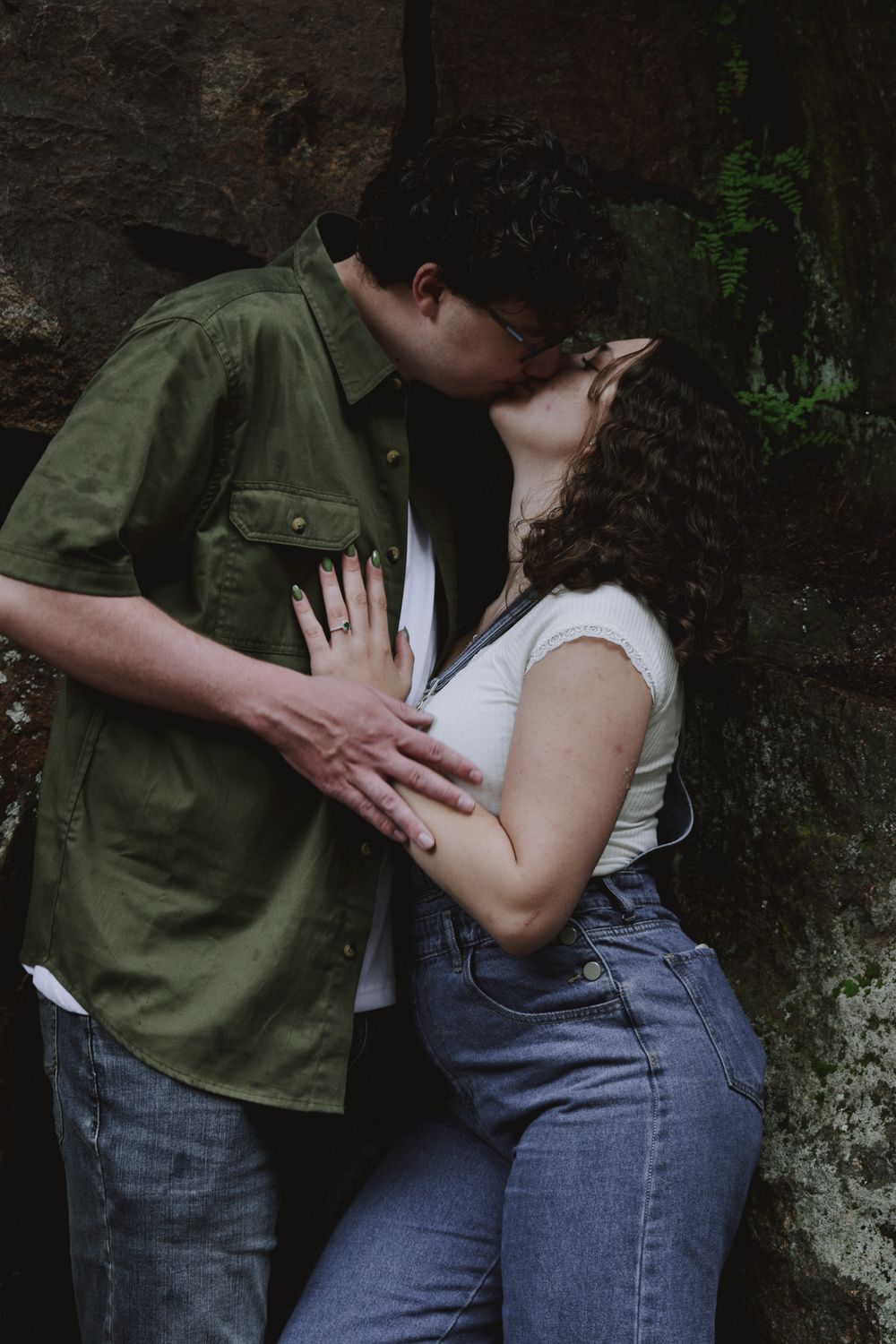 Couple sharing a romantic kiss against a mossy rock wall, woman wearing jeans and white top with engagement ring.