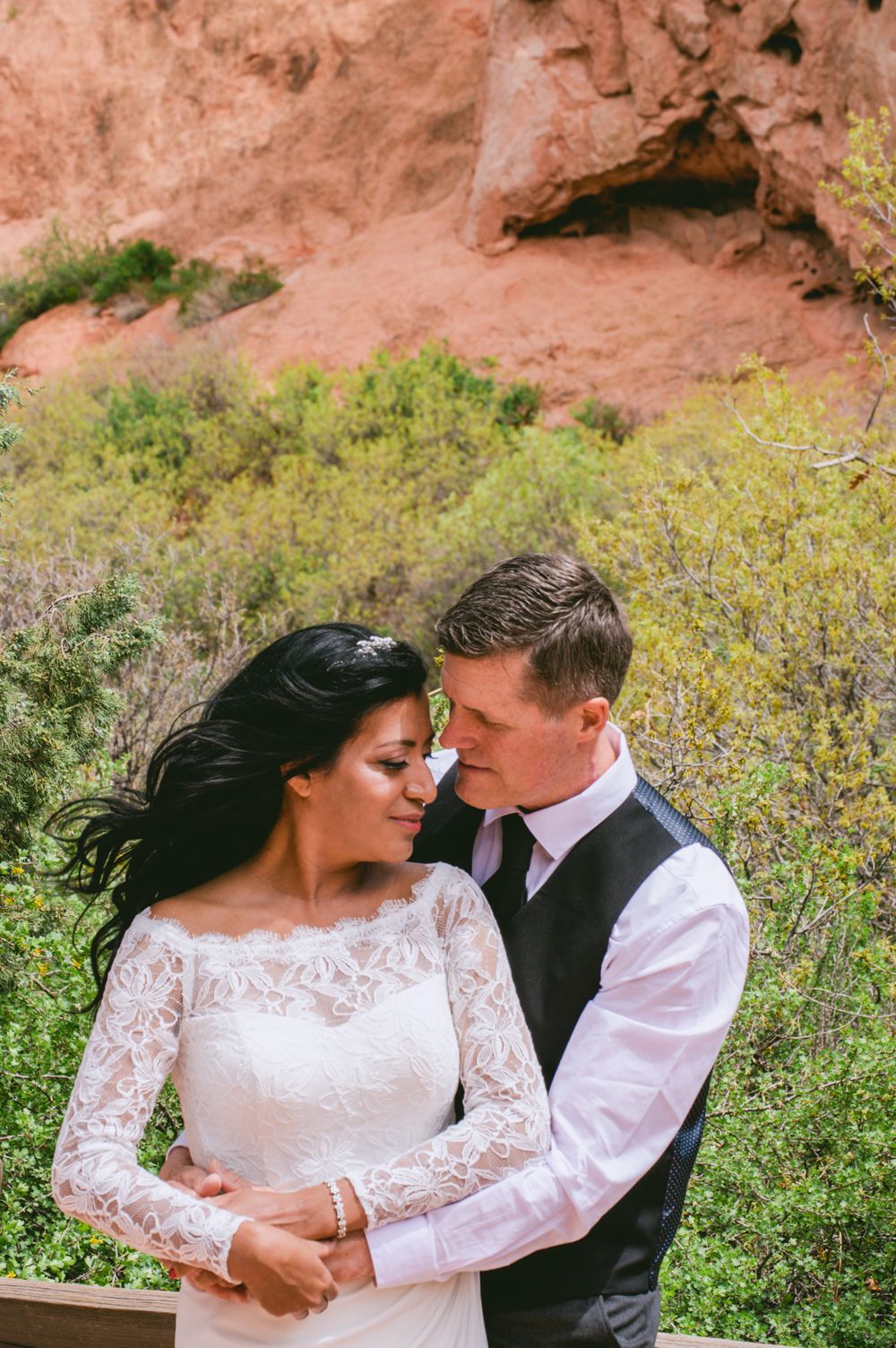 Bride in lace wedding dress and groom in vest embracing tenderly before red rock canyon backdrop.