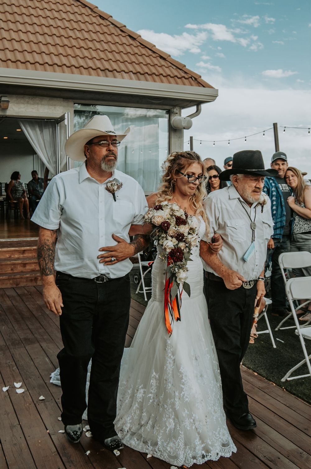 A bride in white lace gown walks the aisle escorted by two men in cowboy hats outdoors.