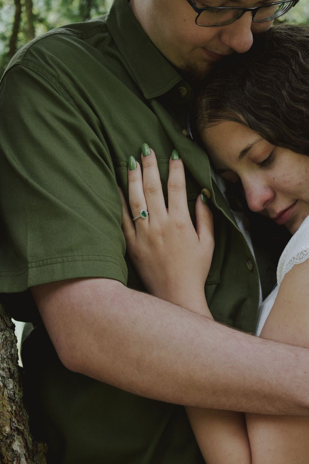 Woman with green nails and engagement ring resting her head on mans chest wearing green shirt outdoors.